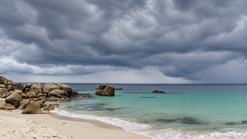 Granite boulders and a curve of sand beside turquoise water beneath grey clouds