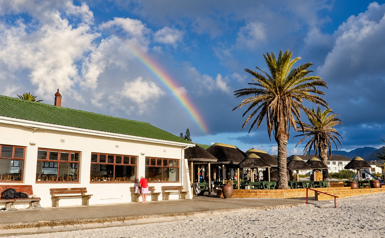 A rainbow above a green-roofed beachfront building and palm trees at Fish Hoek
