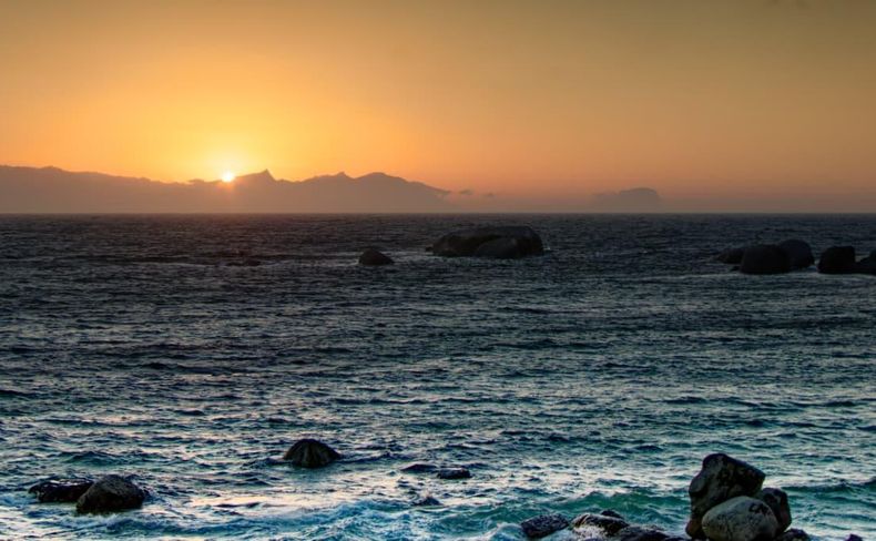 Sunrise above the mountains across False Bay with waves and granite shoreline rocks
