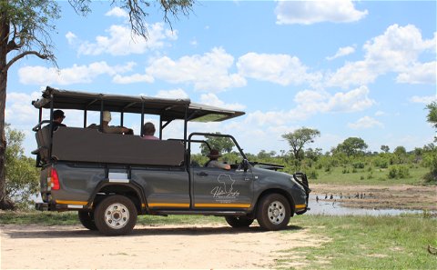 Grey safari vehicle with guests looking out over a dam with birds under blue skies with white clouds