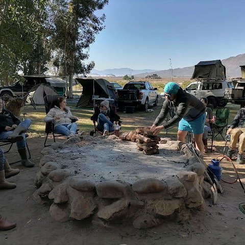 Campers sitting around a fire pit at Wolvenberg Boskamp at Wolvenberg & Stille Waters campsites, while a man is packing and starting a fire.