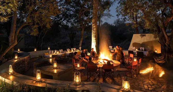 At Simbavati Camp George, guests can enjoy a cozy outdoor atmosphere around a campfire, surrounded by trees. This setting offers the perfect environment for storytelling and sharing experiences under the stars.