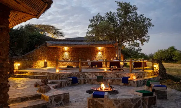 The image features a fire pit surrounded by stones, with a tree nearby. In the background, there is a house or building under a cloudy sky, with some outdoor furniture visible on the ground.