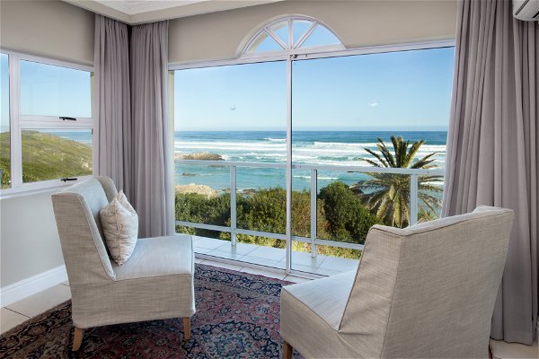 Two white chairs on a Persian carpet inside a white home with wide windows overlooking the Indian Ocean. Palm trees in view and local cape fynbos