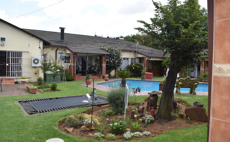 A spacious residential backyard featuring a grassy lawn, an in-ground trampoline, a swimming pool, and a single-story house with a covered patio area.