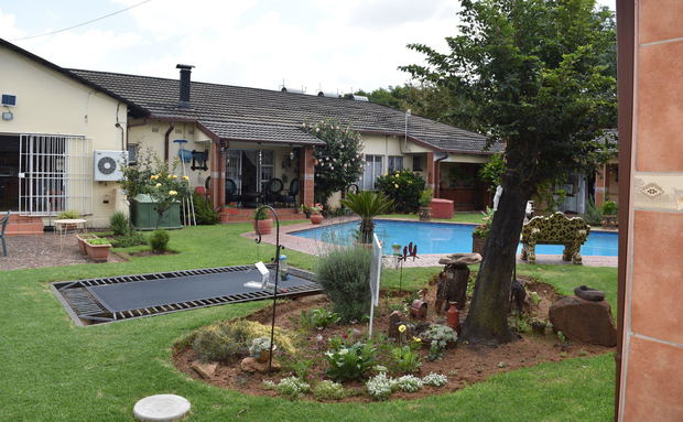 A spacious residential backyard featuring a grassy lawn, an in-ground trampoline, a swimming pool, and a single-story house with a covered patio area.
