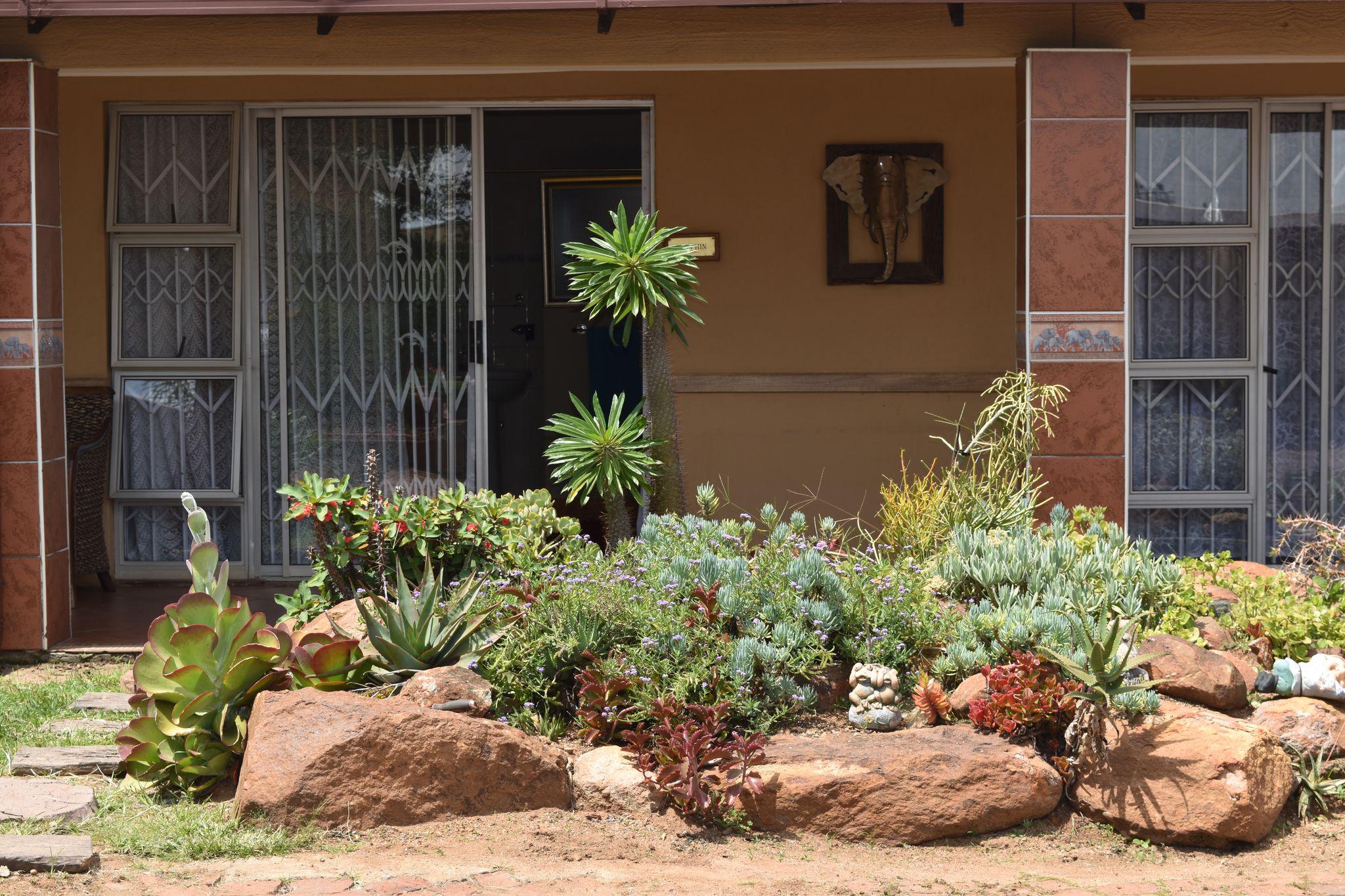 A well-maintained rock garden filled with various succulents and green shrubs stands in front of a house entrance featuring glass sliding doors and security gates