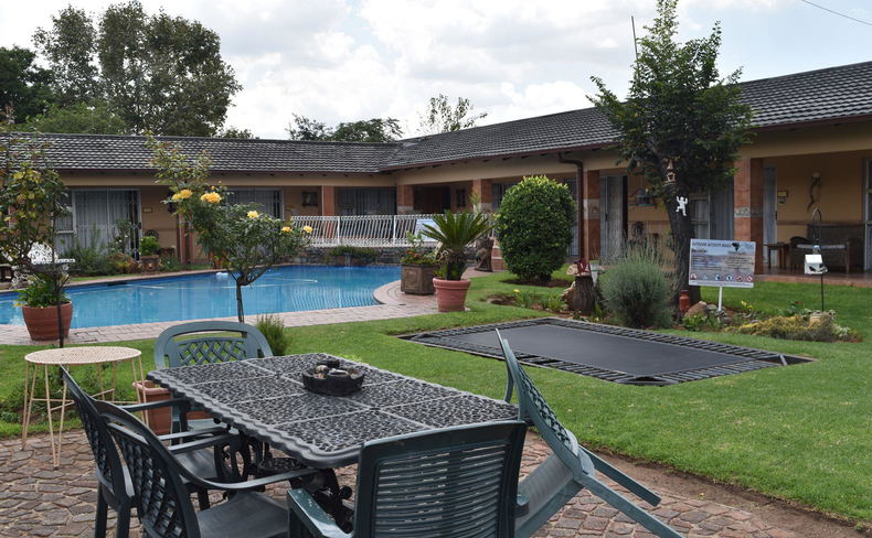 An outdoor patio features a dark-colored dining table and chairs in the foreground, looking out toward a swimming pool, a green lawn, and a single-story brick building structure under an overcast sky An outdoor patio features a dark-colored dining table and chairs in the foreground, looking out toward a swimming pool, a green lawn, and a single-story brick building structure under an overcast sky