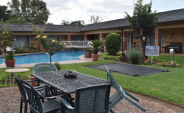 An outdoor patio features a dark-colored dining table and chairs in the foreground, looking out toward a swimming pool, a green lawn, and a single-story brick building structure under an overcast sky An outdoor patio features a dark-colored dining table and chairs in the foreground, looking out toward a swimming pool, a green lawn, and a single-story brick building structure under an overcast sky
