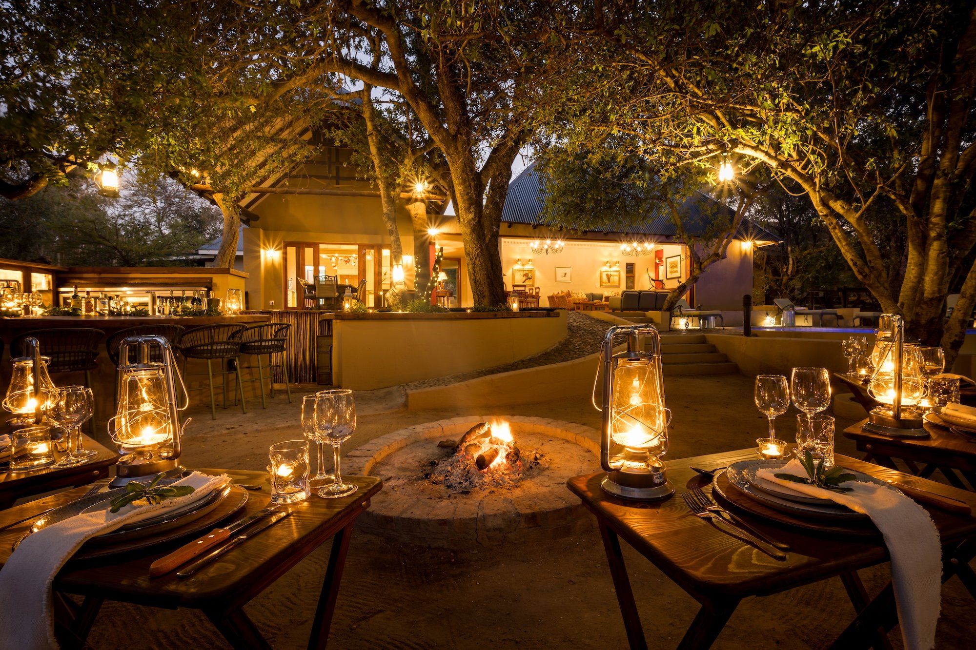 Candlelit outdoor dining tables with lanterns beside a stone fire pit under large trees at dusk, with a lodge in the background.