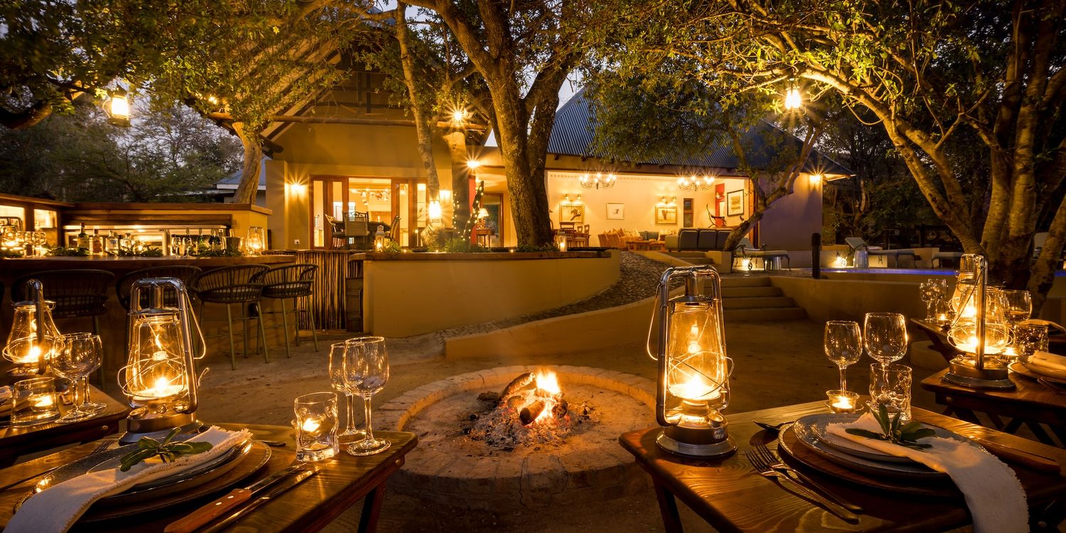 Candlelit outdoor dining tables with lanterns beside a stone fire pit under large trees at dusk, with a lodge in the background.