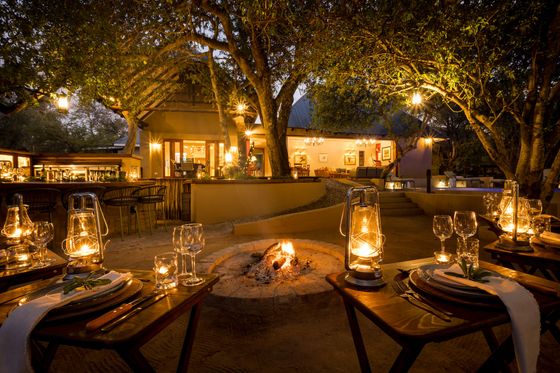 Candlelit outdoor dining tables with lanterns beside a stone fire pit under large trees at dusk, with a lodge in the background.