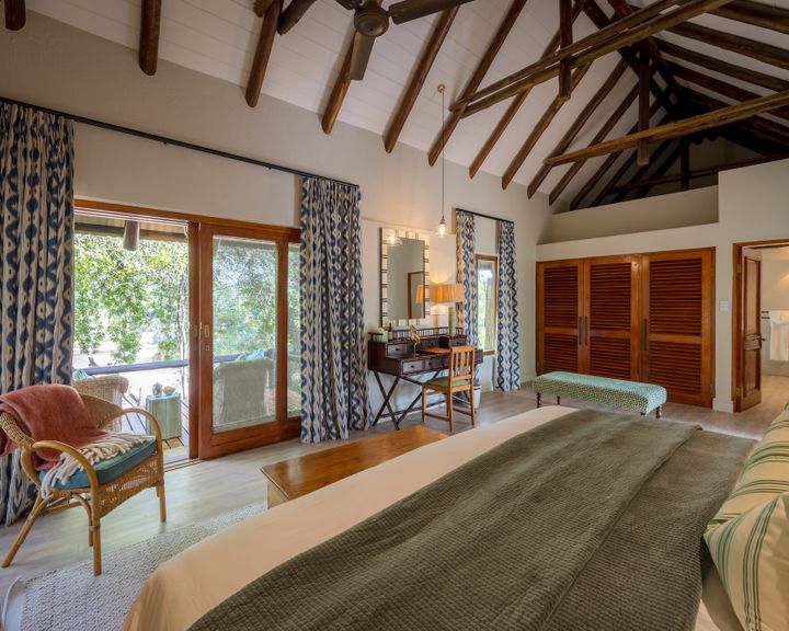 Spacious bedroom with a king bed, vaulted wooden-beamed ceiling, French doors to a deck, and a wicker armchair.