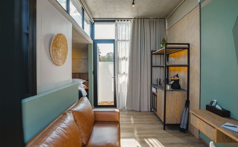 Living area with a tan leather sofa, a woven wall disc, wooden wine shelf with glasses, a desk beside a glass door.