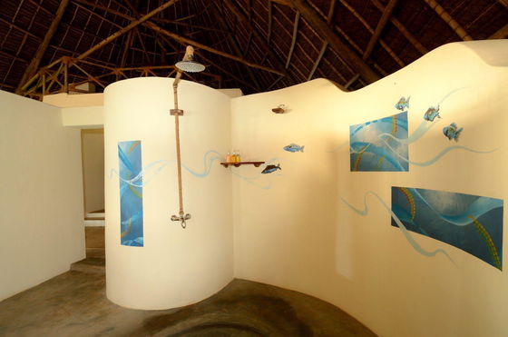 Open-air shower under a high thatched roof at Unguja Lodge Zanzibar, with curved white plaster walls, rope-wrapped overhead shower, concrete floor, small wooden shelf holding toiletries, and blue fish and wave mural panels painted on the walls.