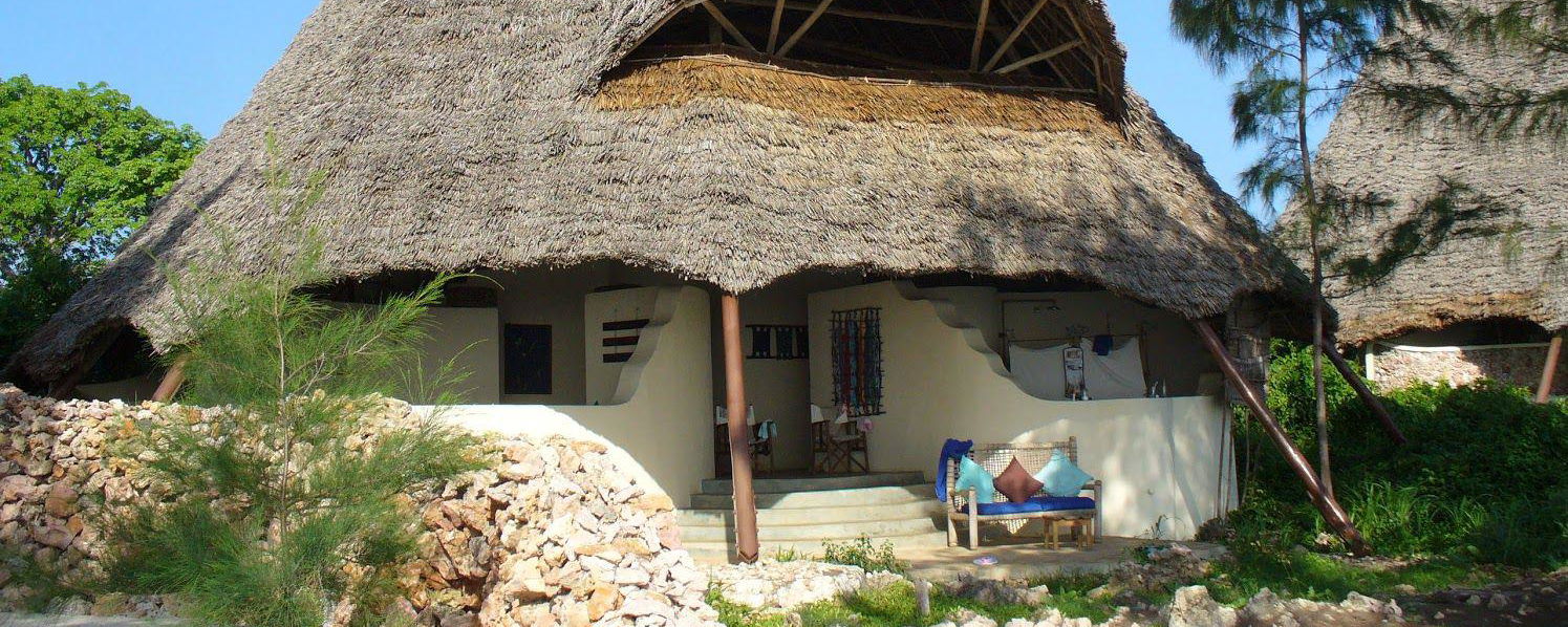 Large conical thatched-roof tropical hut in Zanzibar with twin peaks, white curved stucco lower walls, shaded open porch with blue-cushioned wooden bench, low coral-stone wall and small trees in foreground under clear blue sky