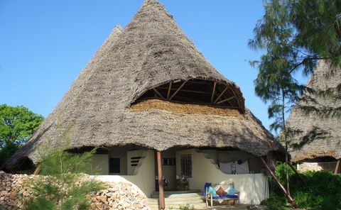 Large conical thatched-roof tropical hut in Zanzibar with twin peaks, white curved stucco lower walls, shaded open porch with blue-cushioned wooden bench, low coral-stone wall and small trees in foreground under clear blue sky