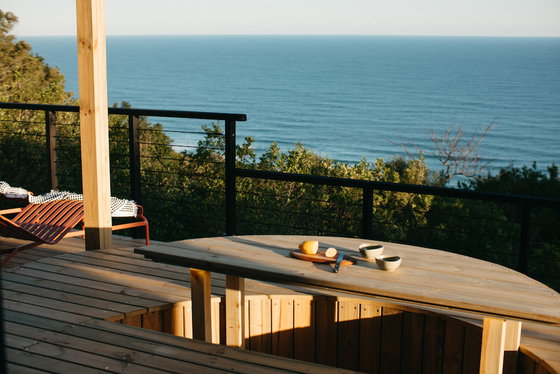 Wooden ocean-view deck with round picnic-style table holding a cutting board with halved lemon and knife and two small bowls, slatted wooden benches, metal railing, red lounge chair with striped blanket, green treeline and calm blue sea on the horizon.