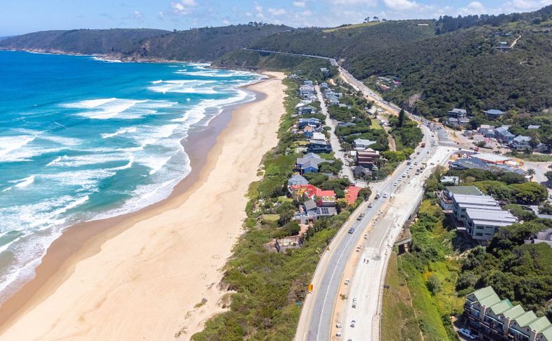 Aerial view of a long sandy beach beside a busy coastal road, with waves breaking on the left and hillside homes on the right.