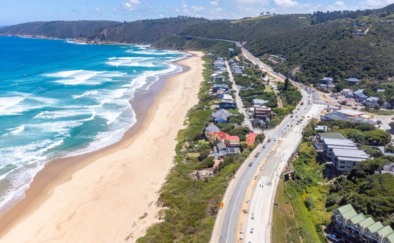 Aerial view of a long sandy beach beside a busy coastal road, with waves breaking on the left and hillside homes on the right.