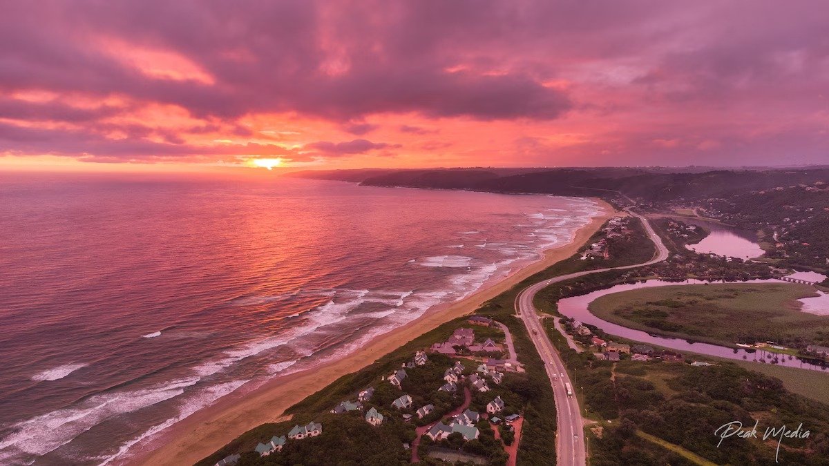 Aerial sunset view of a coastal road, rows of houses and a pink-tinted lagoon beside a long sandy beach.