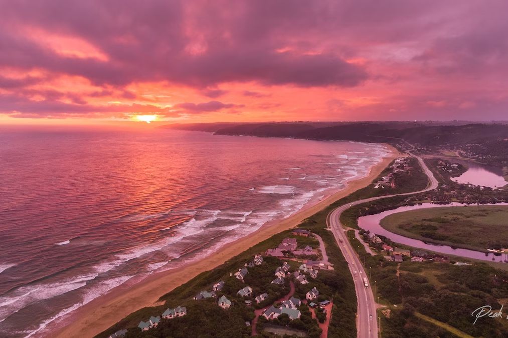 Aerial sunset view of a coastal road, rows of houses and a pink-tinted lagoon beside a long sandy beach.