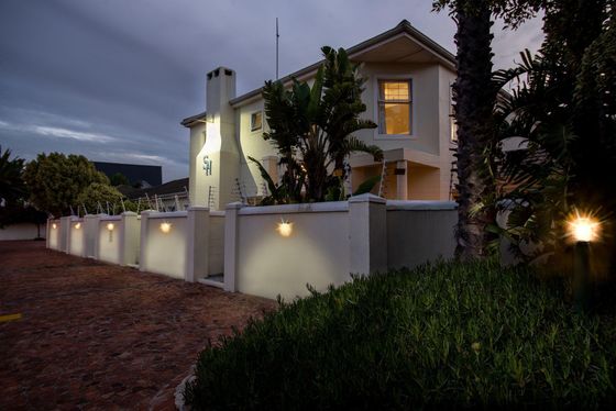 Evening view of a cream-colored guest house behind a white wall lit by small wall lights, with a brick driveway and palm plants.