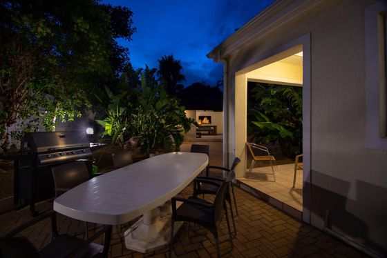 Evening patio with a long white outdoor dining table, dark chairs, a gas barbecue, and a lit fireplace in the garden.