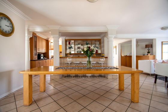 Long wooden dining table with a flower vase, set in a open-plan room with bar stools, a wood kitchen and a lounge area.