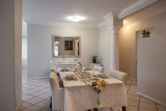 Dining room with a long table set with white cloth, floral runner, bowls, glasses, and cream upholstered chairs.