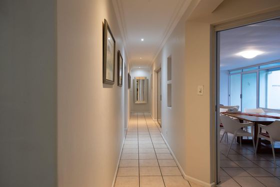 Tiled hallway with framed prints and a mirror, leading to a dining area with a wooden table and white chairs.