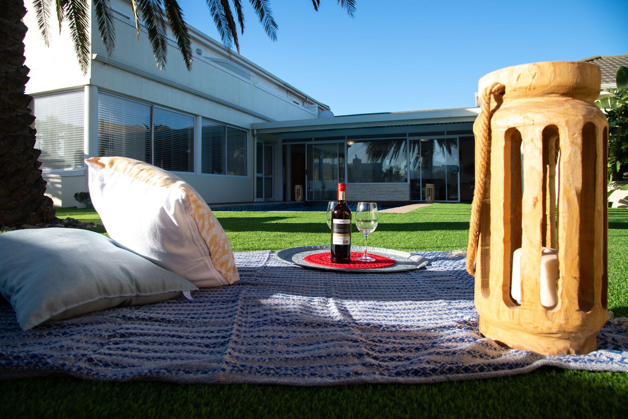 Picnic setup on a blue woven blanket on green lawn, with a bottle of red wine, two glasses, cushions and a wooden lantern.