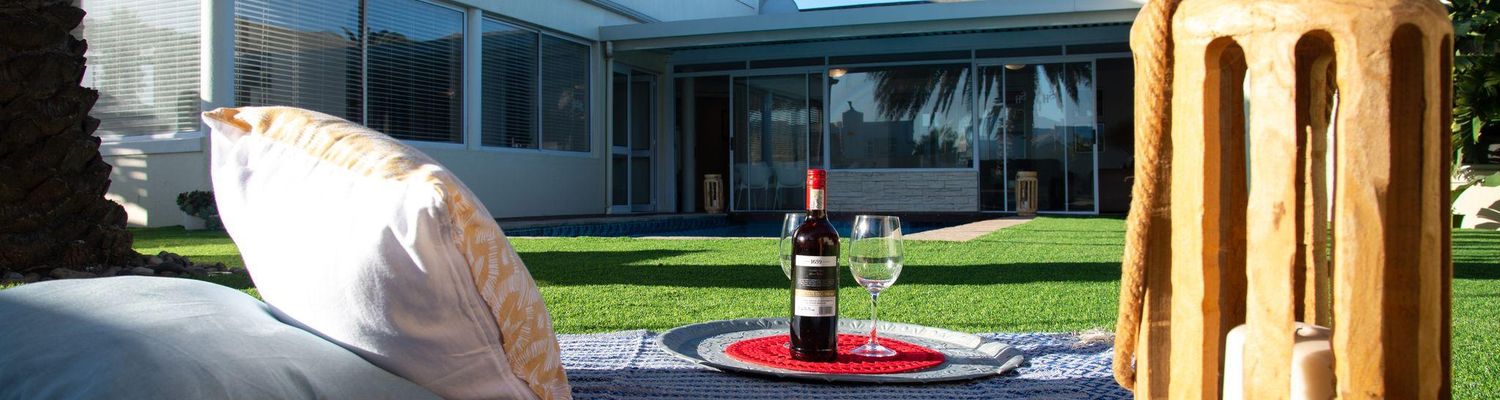 Picnic setup on a blue woven blanket on green lawn, with a bottle of red wine, two glasses, cushions and a wooden lantern.