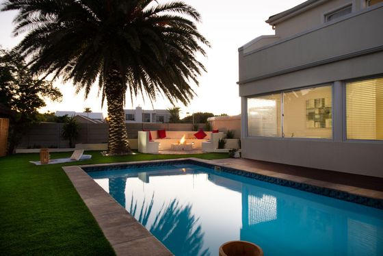 Swimming pool with a palm tree, sunken lounge with red cushions and a fire pit in a garden at dusk.