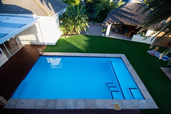 Rectangular swimming pool with tiled edges beside a wooden deck, lawn, and a thatched-roof lounge area.