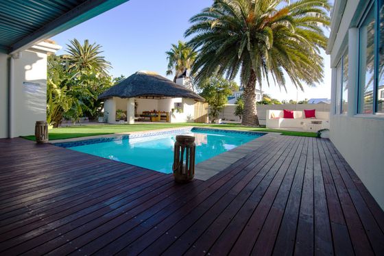 Swimming pool on a timber deck beside a thatched gazebo, with palm trees and red cushioned seating.