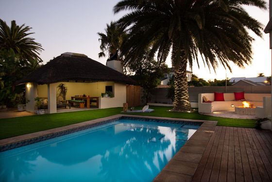 Swimming pool beside a thatched gazebo, lawn, palm tree and red-cushioned lounge seating at dusk.