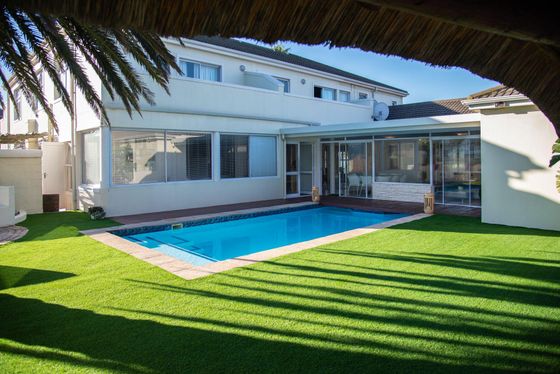 Rectangular swimming pool with green artificial lawn and a white two-storey house with glass sunroom, framed by a thatched roof edge.
