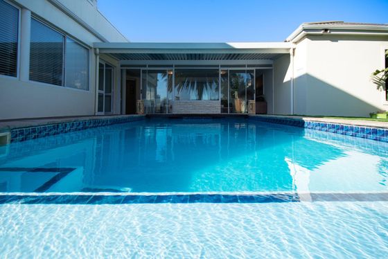 Swimming pool with blue tiled edges in front of a covered glass-walled patio of a white house.