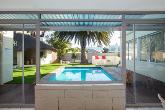 Swimming pool in a sunny garden with a palm tree, white lounge seating with red cushions, and a thatched rondavel beside it.