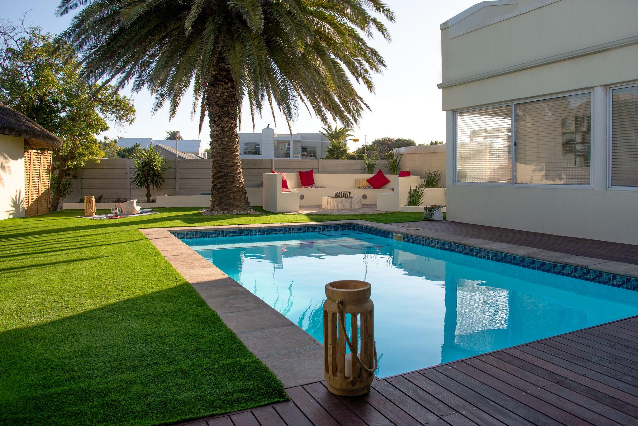 Swimming pool with wooden deck and artificial lawn, beside a tall palm and cream seating area with red cushions.