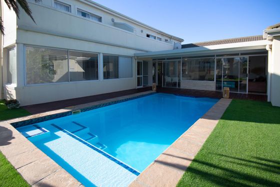 Rectangular swimming pool with a tiled edge beside a double-storey house, with a covered deck and artificial lawn.