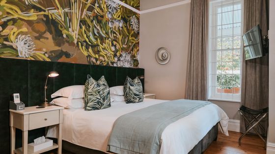 A bedroom with a made-up double bed, green velvet headboard, botanical wall mural, bedside lamp, and wall-mounted TV.