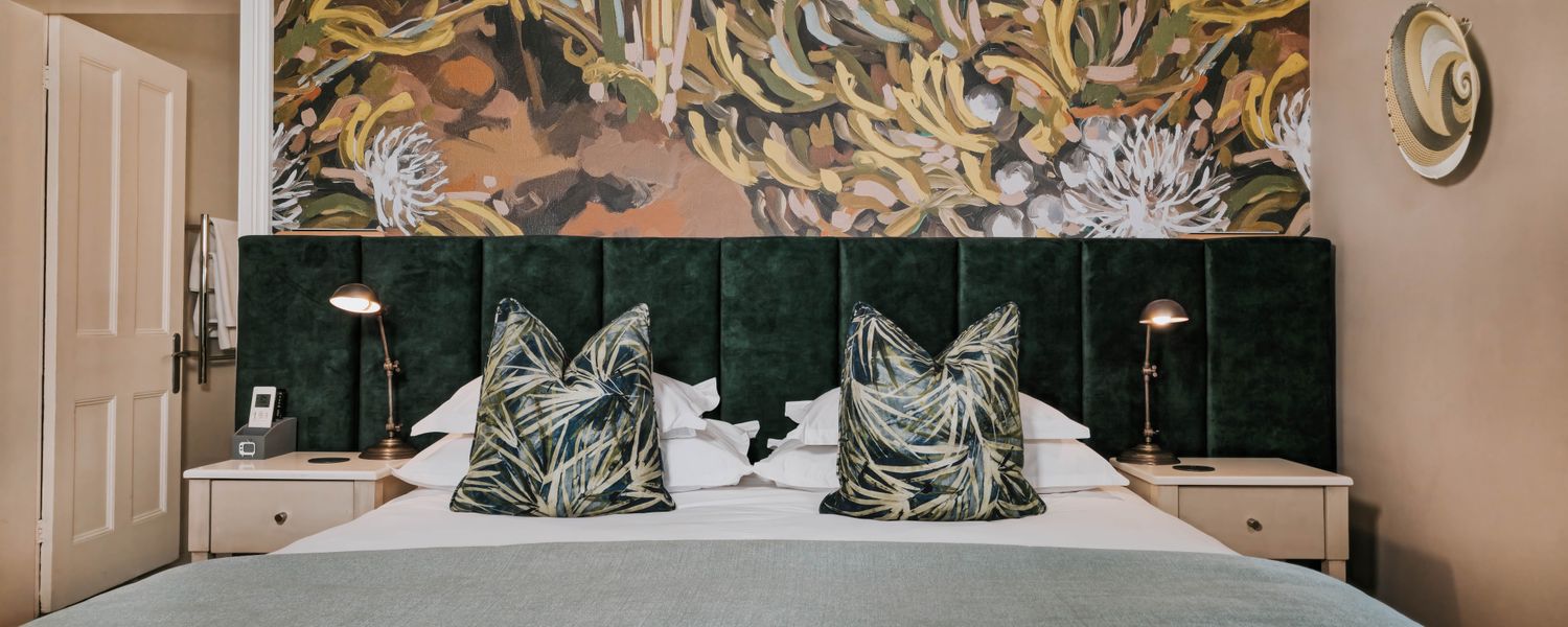 Double bed with dark green velvet headboard, two patterned cushions, and a botanical wall mural behind it.