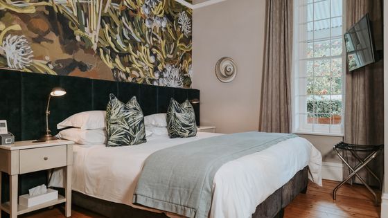 Bedroom with a large white bed, green patterned cushions, a botanical wall mural above a dark green upholstered headboard, and a window with garden view.