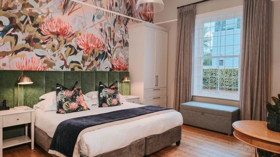 Bedroom with a large bed under a floral botanical wall mural, green upholstered headboard, and a window with grey curtains.