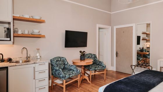 Studio apartment with a kitchenette, two patterned armchairs at a small round wooden table, and a wall-mounted TV.