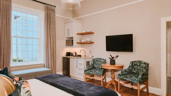 Studio apartment with a made bed, green leaf-print armchairs, a round wooden table, a wall-mounted TV, and a small kitchenette.