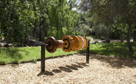 A large interactive wooden sculpture installed along the Stellenbosch Art Mile, surrounded by lush greenery and dappled sunlight. 