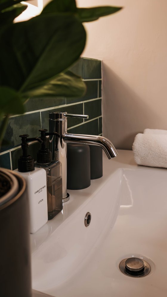 Bathroom sink with chrome tap, dark green tiled splashback, toiletry pump bottles, and a folded white towel beside a leafy plant.